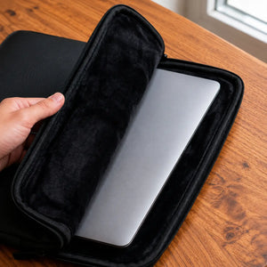 A black zippered laptop sleeve with a plush interior lining, shown open with a silver laptop inside on a wooden table. A hand is holding the sleeve open.